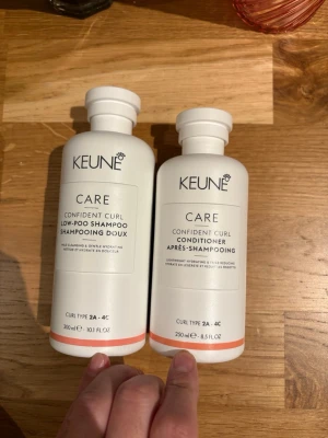 Keune Confident Curl Shampoo & Conditioner - Två vita flaskor från Keune Care-serien: Confident Curl Low-Poo Shampoo (300 ml) och Confident Curl Conditioner (250 ml). Båda är anpassade för lockigt hår (typ 2A-4C) och innehåller essentiella mineraler, kokosolja, sheasmör och vinägerextrakt. Flaskorna har en stilren design med orange detalj.