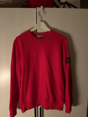 Röd sweatshirt från Stone Island - Helt ny röd sweatshirt från Stone Island med klassisk rund halsringning och ribbade muddar. Tröjan har en ikonisk logotyp-patch på vänster ärm och är tillverkad i mjuk bomull. Perfekt för dig som gillar streetwear och vill sticka ut med färg.