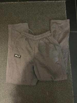Grå mjukisbyxor från PLT - Säljer ett par grå mjukisbyxor från PLT med elastisk midja och snörning. Byxorna har en relaxed passform och ett stilrent PLT-märke på benet. Perfekta för chill dagar och har en mjuk känsla tack vare bomullsmaterialet.