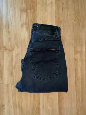 Nudie Jeans  - Säljer ett par svarta jeans från Nudie Jeans. Jeansen har raka ben och hög midja. Materialet är slitstark denim i bomull. Midja 30cm, längd 96cm. JAG SKICKAR EJ FLER BILDER NÄR JAG BÄR JEANSEN! för bättre inblick i passformen rekommenderar jag att googla på modellnamnet. För storleksguide kolla måtten noggrant då jeansen alltid kan vara uppsydda heller krympta och jag ej tar emot returer :) s5,3