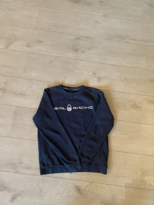 Mörkblå Sail Racing sweatshirt - Mörkblå sweatshirt från Sail Racing med vitt tryck på bröstet och logga i nacken. Klassisk rund halsringning och långa ärmar. Tillverkad i mjuk bomullsmix som är skön att ha på sig. Perfekt för dig som gillar stilren och sportig look.Sail Racing Sweatshirt Tröja Marinblå Vit text Logotyp Casual Sportig Bekväm Vardaglig Mode Stilren Trendig Höst Vinter Vår Herr Dam Unisex Storlek (ange storlek) Bomull Blandning Fleecefodrad Mjuk Varm Premium Kvalitet Design Skandinavisk Segling Båt Hav Friluftsli