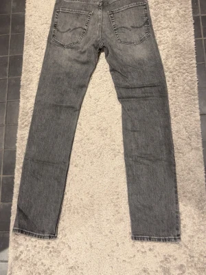 Grå Jack & Jones jeans slim fit 27/32 - Säljer ett par grå jack & Jones jeans med klassisk femficksdesign och raka ben. Jeansen har en snygg tvättad look och är tillverkade i slitstarkt bomullsmaterial. Passformen är slim och midjan är normal. Perfekta till en lite mer classy look.