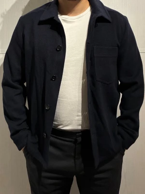 Mörkblå overshirt J.Lindeberg - Stilren mörkblå overshirt från J.Lindeberg i mjuk ull. Klassisk krage, knappstängning framtill och en bröstficka. Perfekt att slänga på över en t-shirt för en clean look. Passformen är normal och materialet känns lyxigt och varmt. Oanvänd. 