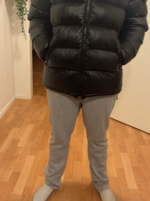 Svart Moncler pufferjacka  - Säljer en svart pufferjacka från Moncler med klassisk logga på ärmen. Jackan har quiltad design, dragkedja framtill och justerbar snörning nedtill. Materialet är glansig polyester och dunfyllning, perfekt för kalla dagar. Insidan har en unik Moncler-serieetikett. Den går att skanna 