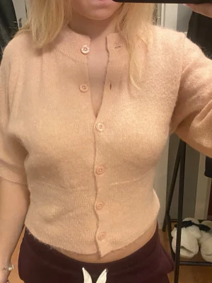 Ljusbeige stickad kofta med kort ärm - Säljer en mjuk och fluffig ljusbeige stickad kofta med korta ärmar och rund hals. Koftan har knappar framtill och är lite croppad i modellen, vilket ger en trendig look. Perfekt att styla med jeans eller kjol för en chill vibe.