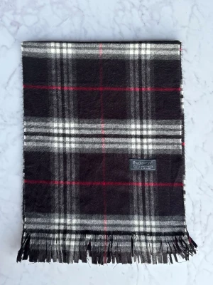 Rutig ullväst från Burberrys - Snygg rutig väst från Burberrys i 100% lammull, tillverkad i England. Västen har klassiskt rutmönster i svart, grått, vitt och röda linjer samt fransad nederkant. Perfekt för dig som gillar tidlös design och vill ha något som sticker ut lite extra.