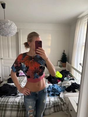 Blommig croppad topp från Adidas - Säljer en färgstark croppad topp från Adidas med stort blommönster i rött, blått, svart och orange. Toppen har korta ärmar och bred rosa kant nertill. Perfekt för dig som gillar sportiga och unika plagg med mycket färg.