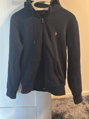 Ralph Lauren zip hoodie - En fräsch zip hoodie ifrån Polo Ralph Lauren. Den är i väldigt bra skick även fast den är använd ett par gånger, ser inprincip ut som ny. Den är mörkblå och i storlek XS. Hör av dig vid funderingar! 