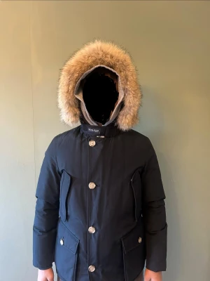 Svart parkajacka från Woolrich med päls - Parkajacka från Woolrich i svart med stor huva kantad av beige päls. Jackan har långa ärmar, stora fickor med lock och knappar framtill. Materialet är polyester och jackan är fodrad för extra värme. Perfekt för kalla vinterdagar.(äkta päls)