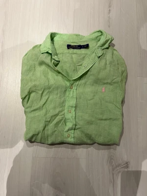 Grön linneskjorta från Polo Ralph Lauren - Fräsch grön skjorta från Polo Ralph Lauren i mjukt linne. Klassisk krage, knappar framtill och en liten rosa logga broderad på bröstet. Perfekt för dig som gillar stilrena och färgglada plagg. Skjortan har en avslappnad passform och är lätt att matcha.