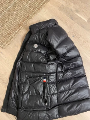 Svart dunväst från Moncler - Säljer en svart dunväst från Moncler med quiltad design och glansig finish. Västen har dragkedja framtill, fickor med dragkedja och en snygg röd-vit-blå rand längs dragkedjan. Perfekt för lager-på-lager och har en modern, loose passform. Pris går att diskutera. Storleken är för lite större personer långa och folk mer lite mer fett elle muskler. Västen är äkta med Scan 