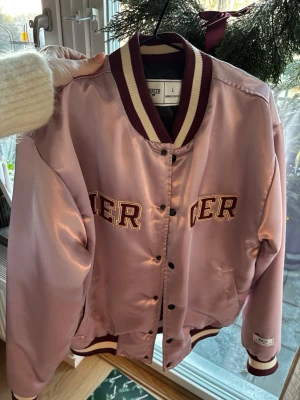 MERCER - satin bomberjacka - Säljer en riktigt snygg bomberjacka från Mercer i satin. Jackan har vinröda och vita ribbade muddar, tryckknappar framtill och broderade bokstäver på bröstet. Perfekt statement-plagg med cool retrovibe och stora fickor. Egenskaper//  Vindtät Vattentät Om produkten Material: glossy satin Yttermaterial: premium chenille branding Detta är en Unisexprodukt Maskintvätt i högst 40˚C Använd ej blekmedel Ska ej torktumlas Strykning, högsta temperatur 150 °C Kemtvätt