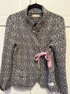 Kofta från Odd Molly - Säljer en svartvit mönstrad kofta från Odd Molly med lång ärm och snygg krage. Koftan har en ljusrosa rosett i sidan och är stickad i mjukt material. 
