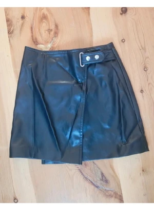 Svart omlottkjol i skinnimitation - Säljer en svart kort omlottkjol i skinnimitation. Kjolen har en snygg detalj med bälte och silverfärgade knappar i midjan. Perfekt för dig som gillar en edgy och trendig look.