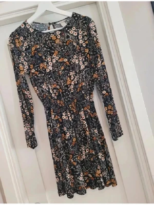 Blommig kortklänning från H&M - Supersöt kortklänning från H&M med blommigt mönster i svart, vit och gul. Klänningen har långärmade ärmar, rund hals och ett mjukt, följsamt material som sitter bekvämt. Perfekt för dig som gillar färg och mönster!
