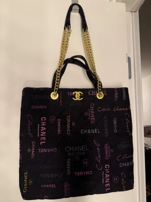 Svart märkes axelväska med kedja - Säljer en svart axelväska med quiltad look och färgglada logotexter i rosa, lila och beige. Väskan har guldiga kedjehandtag och klassisk CC-detalj framtill. Insidan är fodrad i svart tyg med innerfack och dragkedja. Perfekt för dig som vill sticka ut. Aldrig använt