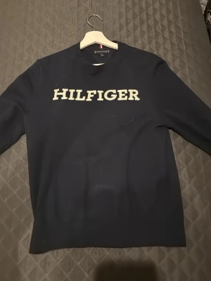 Mörkblå tröja Tommy Hilfiger - Snygg mörkblå tröja från Tommy Hilfiger med stort vitt Hilfiger-tryck på bröstet. Tröjan har rund hals med ribbad kant och långa ärmar. Perfekt för dig som gillar klassisk stil med en modern touch.