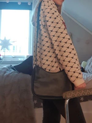 Svart axelväska i skinnimitation - Säljer en stilren svart axelväska från Even&Odd med justerbar rem och dragkedja upptill. Väskan har en enkel, mjukt rundad form och är gjord i skinnimitation. Perfekt storlek för mobil, plånbok och nycklar. Remmen har diskret logga invävd.