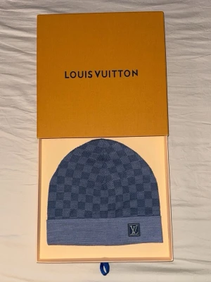 Blå rutig mössa från Louis Vuitton - Säljer en snygg blå mössa från Louis Vuitton med klassiskt rutigt mönster och LV-logga framtill. Använd 3-4 gånger