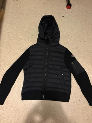 Svart Moncler pufferjacka med huva - Svart pufferjacka från Moncler med stickade ärmar och huva. Jackan har ribbade muddar, dragkedja framtill och fickor med knappar. Moncler-logga på ena ärmen och en extra ficka med dragkedja. Perfekt för dig som vill ha en snygg och varm jacka med streetkänsla.