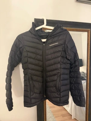 Svart dunjacka Peak Performance XS - Svart dunjacka från Peak Performance i storlek XS. Jackan har huva, dragkedja framtill och två fickor med dragkedja. Quiltad design och diskret logga på bröstet. Perfekt för kyliga dagar och enkel att matcha med allt.Små defekter (se bild) men inget som märks av. 