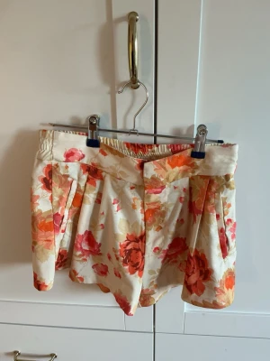 Blommiga shorts från INC Woman - Säljer ett par blommiga shorts från INC Woman med elastisk midja och mjukt tyg. Färgen är krämvit med stora blommor i orange, rosa och rött. Modellen är loose fit och har fina veck framtill för extra stilpoäng.