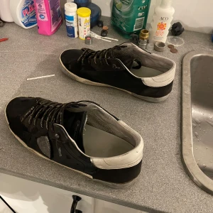 Svarta Philippe Model sneakers med vit häl - Säljer ett par svarta Philippe Model sneakers med vit häl och platt sula. Skorna har snörning med svarta och vita skosnören där märket står skrivet. Materialet är en mix av skinn och mocka, med rund tå och diskret logga på sidan. Perfekt för dig som gillar stilrena och exklusiva sneakers.