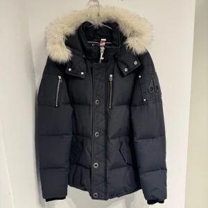 Moose Knuckles marinblå jacka, storlek M - The jacket is recently dry cleaned.  Marinblå Moose Knuckles dunjacka med dragkedja och knappar fram. Jackan flera fickor med dragkedja och tryckknappar samt en logotyp på vänster ärm.