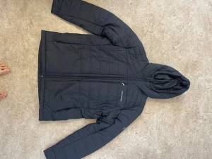 Svart pufferjacka från Peak Performance - Säljer en svart pufferjacka från Peak Performance med huva och snygga quiltade sömmar. Jackan har dragkedja framtill och en praktisk bröstficka med zip. Perfekt för kalla dagar och har en clean, sportig look.