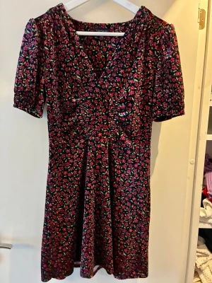 Blommig kortklänning från Topshop - Säljer en söt kortklänning från Topshop med blommigt mönster i rosa och grönt mot svart bakgrund. Klänningen har v-ringning, korta småpuffiga ärmar och lätt rynkade detaljer framtill. Materialet är mjukt och känns som polyester/sammet. Den är ca 82- 84 cm lång 