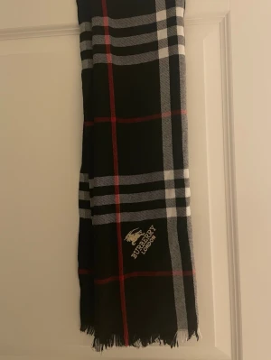Rutig väst från Burberry London - Snygg rutig väst från Burberry London med klassiskt svart, vitt och rött mönster. Västen har fransad nederkant och tydlig Burberry-logga broderad framtill. Perfekt för dig som vill ha en tidlös och trendig look.