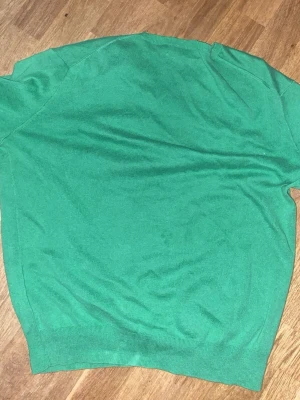 Grön stickad tröja Polo Ralph Lauren - Snygg grön stickad tröja från Polo Ralph Lauren i storlek S. Tröjan har v-ringad hals och korta ärmar, med den klassiska lilla Polo-loggan broderad i lila på bröstet. Perfekt för dig som gillar stilrena och färgglada plagg.