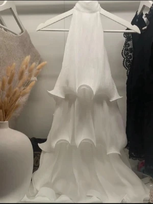 Vit klänning med volanger - Säljer en superfin vit klänning med flera lager volanger som ger en riktigt drömmig och flowy känsla. Klänningen är ärmlös och har en halterneck-design som gör den extra elegant. Perfekt för dig som vill sticka ut med en unik och romantisk look. Använd endast 1 gång!