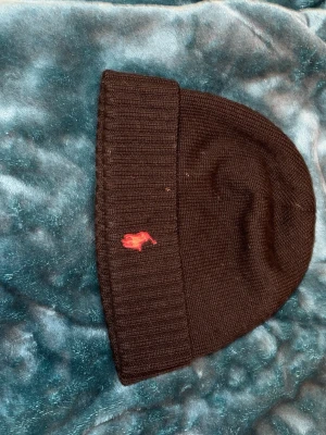 Svart stickad mössa från Polo Ralph Lauren - Svart stickad mössa från Polo Ralph Lauren med klassisk röd broderad logga framtill. Mössan har ribbad kant och är mjuk och skön, perfekt för kyliga dagar. Enkel och stilren design som passar till det. Passar typ alla och sitter still rent på dom flesta