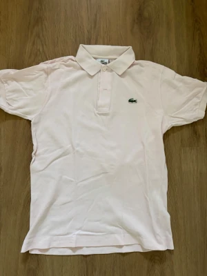 Lacoste piké  - Säljer en ljusrosa piké från Lacoste med klassisk krage och två knappar framtill. T-shirten har korta ärmar och det ikoniska gröna krokodilmärket broderat på bröstet. Materialet är mjuk bomull som känns skönt mot huden. Storleken är S och skicket är mycket bra.                                                                Skriv om ni har några frågor!😇