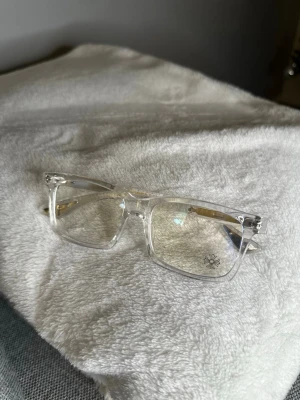 Bluellight chromehearts glasögon  - Säljer ett par stilrena glasögon med transparent båge och rektangulär form. På skalmen finns en snygg blommig detalj i silver som ger extra edge. Linsen har en liten blomma tryckt i hörnet. Perfekt för dig som gillar cleana och moderna accessoarer.