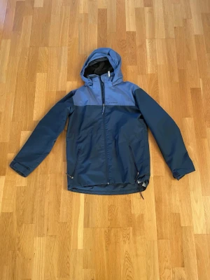 Blå vindjacka från Everest med huva - Snygg tvåtonad blå vindjacka från Everest med huva och hel dragkedja framtill. Jackan har två sidofickor med dragkedja och justerbar nederkant. Perfekt för blåsiga dagar och outdoor-aktiviteter. Meshfoder på insidan för extra komfort.