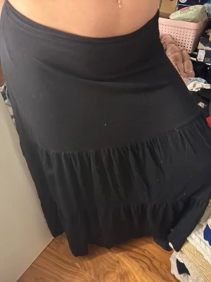 Svart långkjol med volang - Säljer en svart långkjol med bred midja och volangdetaljer. Kjolen har en mjuk och flowy känsla, perfekt för dig som gillar en chill och avslappnad stil. Materialet känns som bomull och kjolen är enfärgad utan mönster.obs finns 3 st jätte jätte små hål