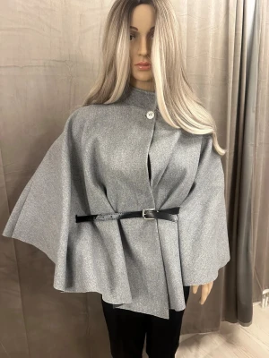 Grå cape-kappa med bälte - Snygg grå cape-kappa med hög krage och dekorativ knapp upptill. Kappan har ett svart smalt bälte i midjan som ger en stilren siluett. Tillverkad i ett mjukt ull-liknande material och har en loose passform med vida ärmar. Perfekt för dig som vill ha en trendig och elegant look.