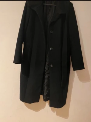 Svart kappa från Lindex, strl 42 - Svart kappa från Lindex i klassisk rak modell med knappar framtill och bälte i midjan. Långärmad och fodrad insida för extra komfort. Perfekt för kyliga dagar och enkel att matcha med olika outfits.