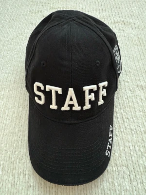 Svart STAFF keps från Balenciaga - Säljer en svart keps från Balenciaga med stor vit STAFF-text broderad framtill och Balenciaga-logga längs insidan och kanten. Kepsen är tillverkad i bomull och har klassisk form med böjd skärm och justerbar rem baktill. Perfekt för dig som gillar streetwear och designer.