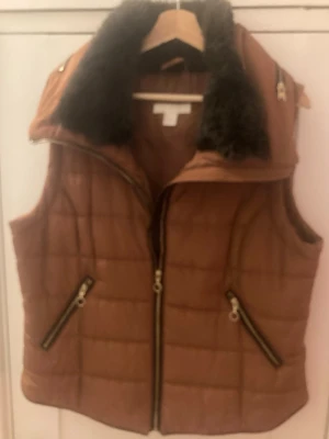 Brun quiltad dunväst med päls - Säljer en brun quiltad dunväst från H&M i storlek L. Västen har en fluffig svart fuskpäls på kragen, två snedställda fickor med dragkedja och en kraftig dragkedja framtill. Perfekt för lager-på-lager under kyliga dagar.