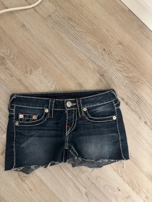 True Religion jeansshorts ’kolla sista bild’ - Säljer ett par mörkblå jeansshorts från True Religion med grova vita sömmar och röda detaljer. Shortsen har låg midja, fem fickor och rå, fransig kant nedtill. Baksidan har märkets ikoniska broderade fickor och patch. Perfekt för dig som gillar street och denim. Några trådar har lossnat men det borde vara lätt att fixa 💞☺️w24, kan byta mot w25 ☺️