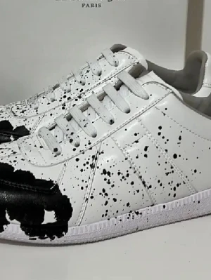 Vit-svarta sneakers från Maison Margiela - Unika vita sneakers från Maison Margiela med svart färgstänk och konstnärlig design. Skorna har snörning, rund tå och är tillverkade i skinn. Perfekta för dig som vill sticka ut med en kreativ och edgy stil.