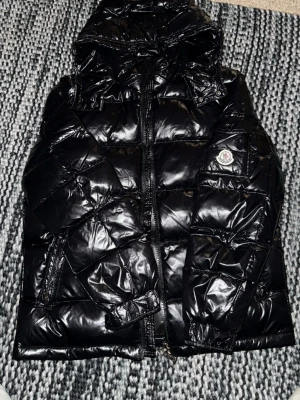 Svart Maja Moncler pufferjacka med huva - Säljer en svart pufferjacka från Moncler med glansig finish och huva. Jackan har dragkedja framtill, två sidofickor med dragkedja och Moncler-logga på ärmen. Perfekt för kalla dagar och har en riktigt snygg streetstil.