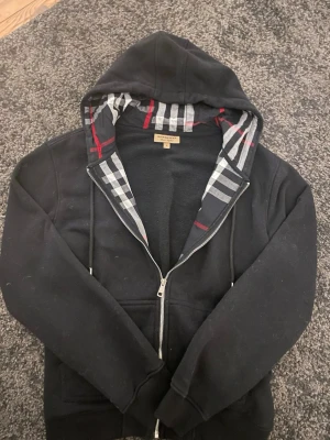 Svart Burberry Zip up - Tröjan är bara använd två gånger och är fortfarande i toppskick. Märkt storlek L men passar mer som en M. Ser i princip ut som ny, inga skador eller slitage. Perfekt tröja i riktigt fint skick. Priset kan diskuteras. Kan eventuellt också byta.