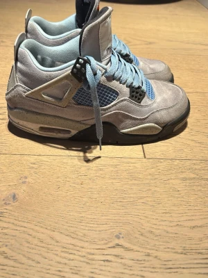 Nike Jordan 4 University Blue sneakers - Nike Jordan 4 University Blue sneakers med ljusblå mocka, svarta och vita detaljer samt specklad sula. Skorna har snörning, meshpaneler och Jumpman-logga på hälen. Perfekt för dig som vill sticka ut med en ikonisk och sportig look.