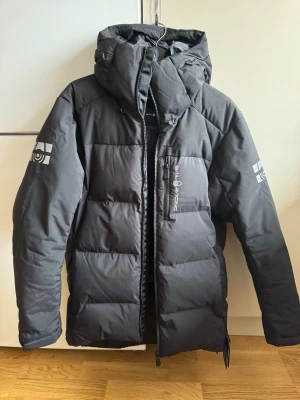 Sail Racing cape down jkt 2.0 - Säljer en svart dunjacka från Sail Racing med 500 FP Allied Feather + Down. Jackan har huva, dragkedja framtill och logga på ärmen. Perfekt för kalla dagar och har en clean, sportig look. Flera praktiska fickor och snyggt puffad design.