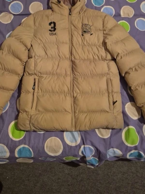 Beige pufferjacka Vinson Polo Club M - Säljer en beige pufferjacka från Vinson Polo Club i storlek M. Jackan har huva, dragkedja framtill och två sidofickor med dragkedja. Coolt tryck med '3 USA' och Polo Club-logga på bröstet. Perfekt för kalla dagar och riktigt snygg stil. Pris kan diskuteras vid snabb affär