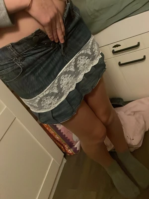 Jeanskjol med spets och plisserad kant - Säljer en cool kort jeanskjol med vit spetsdetalj och plisserad nederkant. Kjolen har klassiska jeansfickor, bälteshällor och en unik patch med döskalle bak. Perfekt för dig som vill sticka ut med en edgy och söt stil.